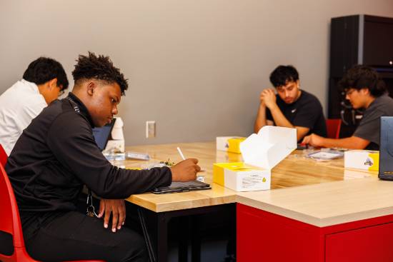 Four people work together around a wooden table covered with laptops, papers, tools, and open white-and-yellow boxes.