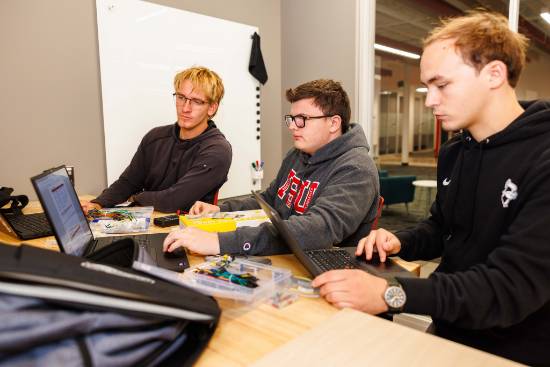 Three students work with laptops and electronic components at a table in a classroom.