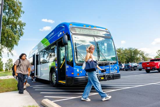 Blue-and-green city bus marked “CAMPUS EAST” stopped in a parking lot as a person walks in front of it making a peace sign.